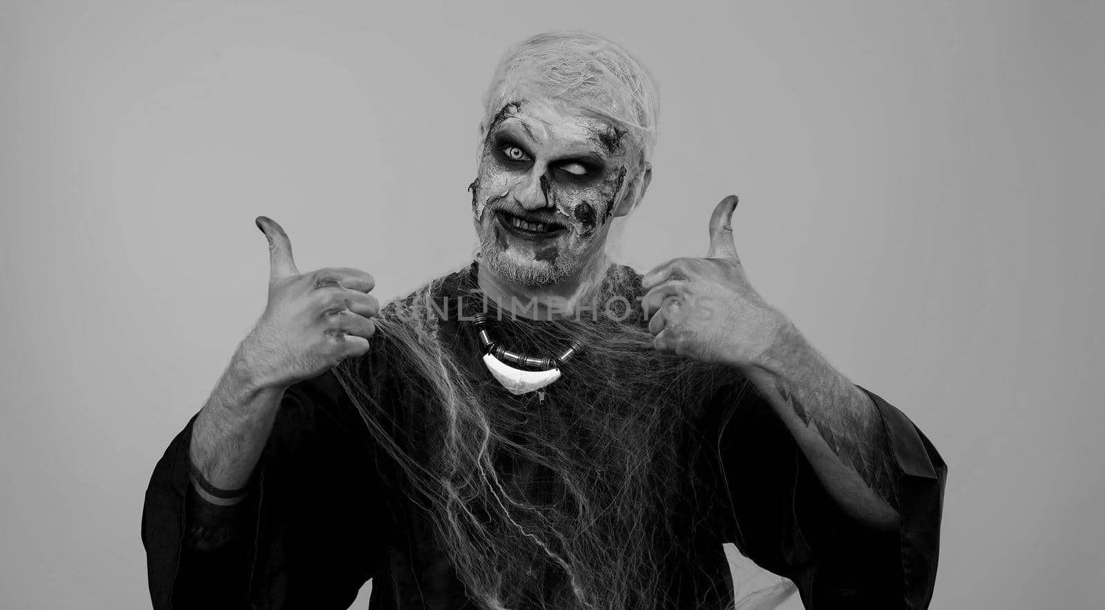 Sinister man with scary Halloween zombie makeup in costume raises thumbs up agrees with something or gives positive reply recommends advertisement likes good. Undead guy with wounded bloody scars face