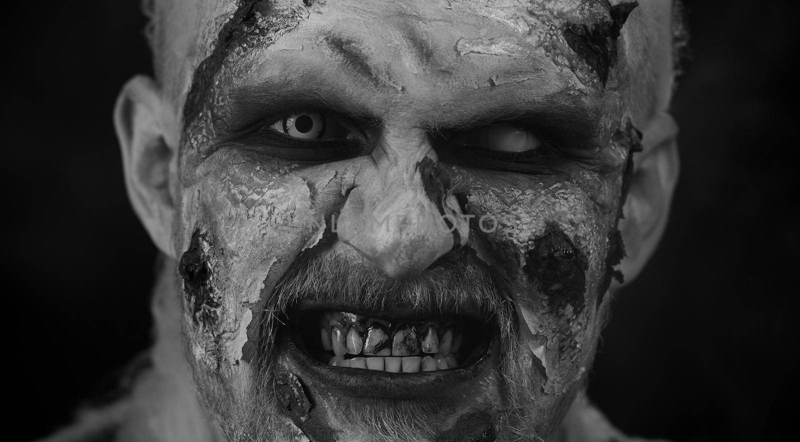 Creepy man close-up face with bloody scars, Halloween stylish zombie makeup. Scary wounded undead guy blows smoke from nose, smiles terribly. Voodoo rituals. Sinister beast, monster, vampire