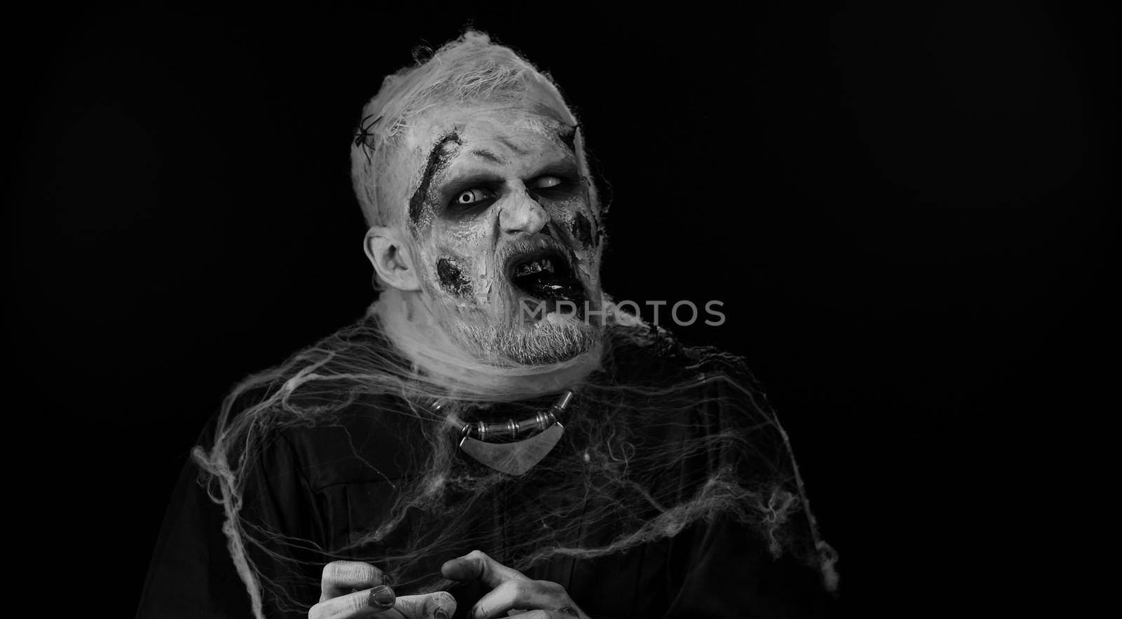 Scary man with horrible scary Halloween zombie makeup in convulsions making faces, looking ominous, trying to scare. Dead guy with wounded bloody scars face isolated against black wall background