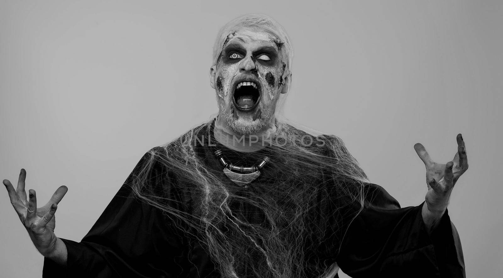 Sinister man in carnival costume of Halloween crazy zombie with bloody wounded scars face screaming, shouting, trying to scare on gray. Horror theme of cosplay wounded undead, beast, monster