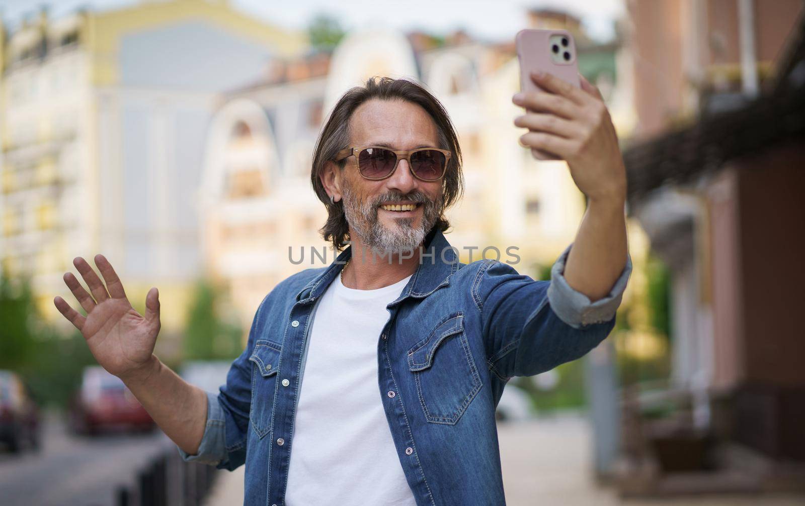 Stylish sexy mature grey bearded man smiling on camera old town wearing casual. Happy senior man on street of European city making selfie using mobile phone. Travel concept. Business on the go.