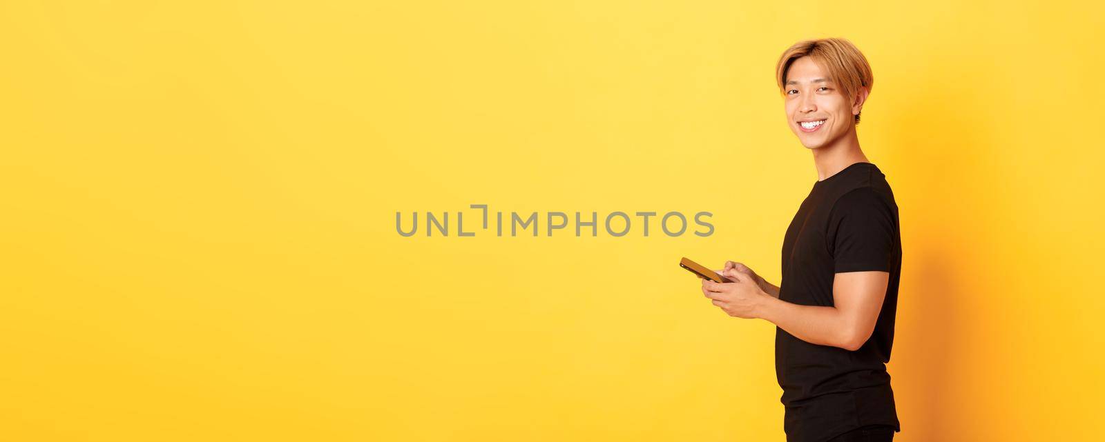 Portrait of handsome stylish asian guy in black outfit, using mobile phone and turning head at camera with satisfied smile, yellow background.