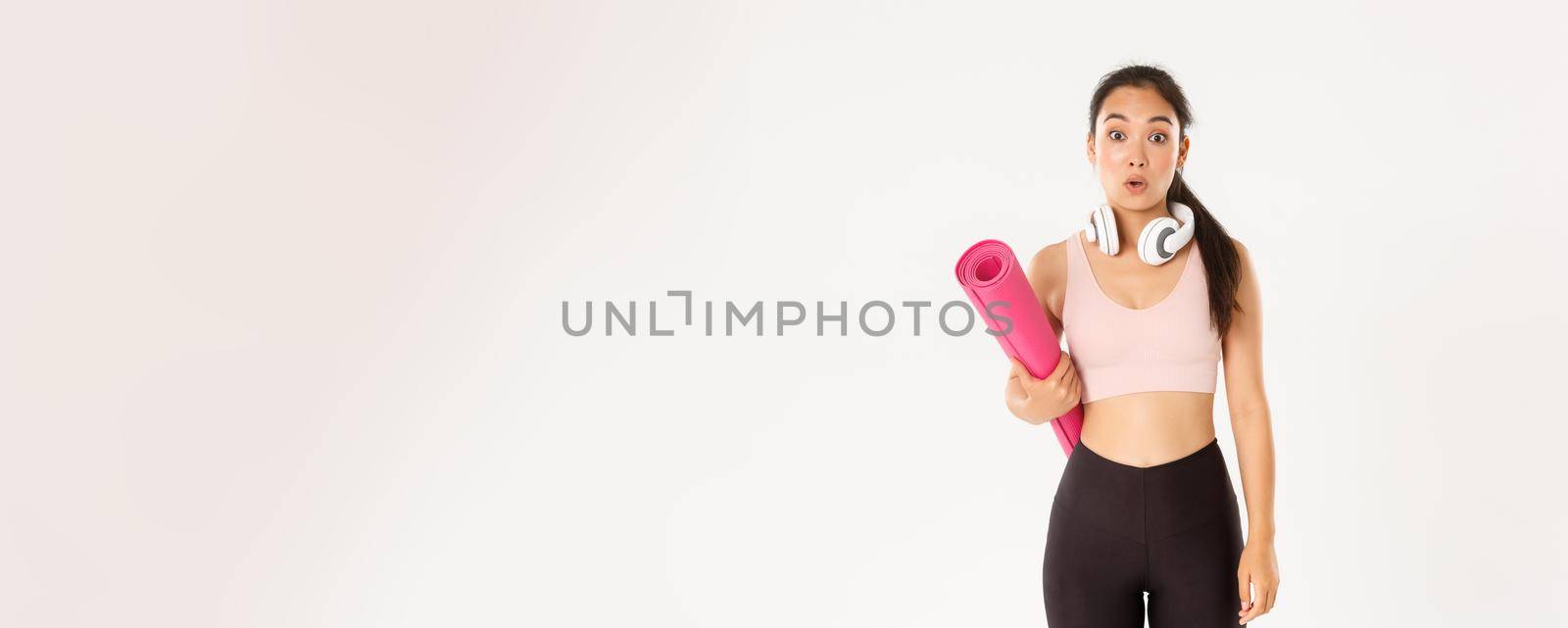 Sport, wellbeing and active lifestyle concept. Confused and shocked asian fitness girl, cant understand why gym closed, holding rubber mat and looking surprised at camera, white background.