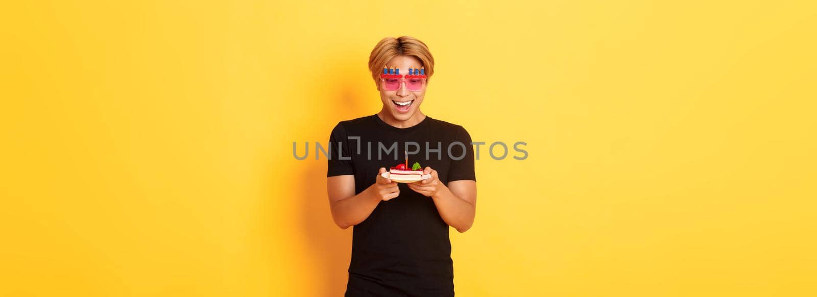 Excited handsome asian guy in party glasses, looking hopeful at birthday cake as celebrating b-day, making wish on lit candle, standing yellow background.