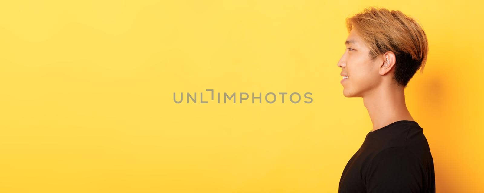 Profile of stylish handsome asian guy with fair hair looking left and smiling, standing over yellow background by Benzoix