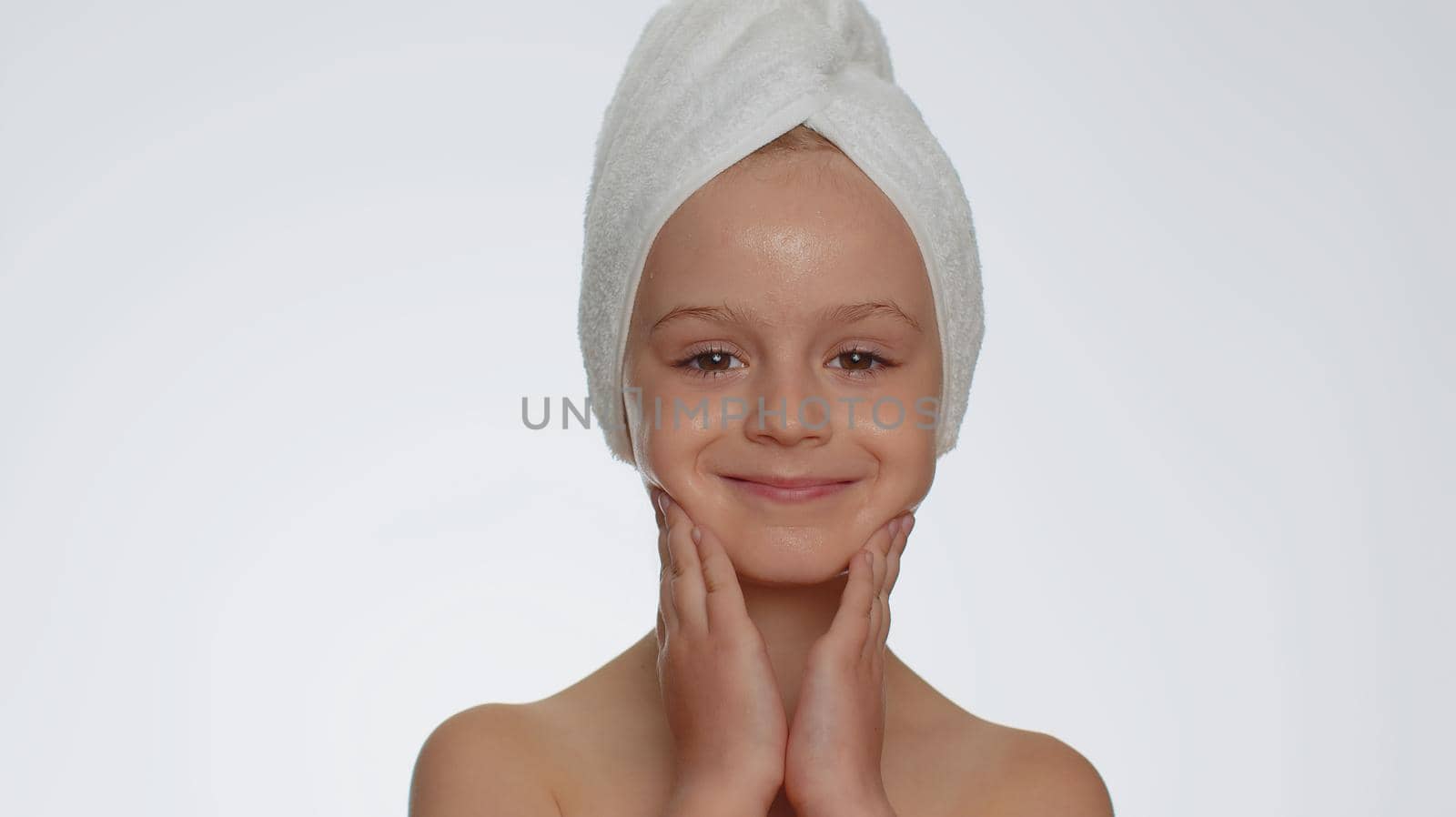 Lovely young child girl after bath in towel on head applying cleansing moisturizing cream. Teenager kid face skincare healthy treatment, natural creme cosmetics. Female portrait. Perfect fresh clean