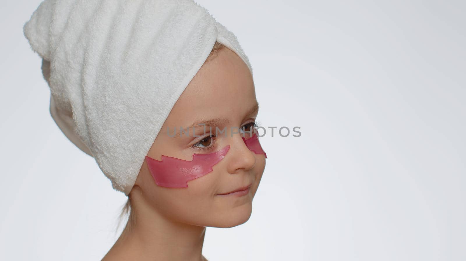 Beautiful young smiling child girl in towel on head applying pink cosmetic patches under eyes. Teenager kid face skin care treatment. Female portrait on white background. Perfect natural fresh clean