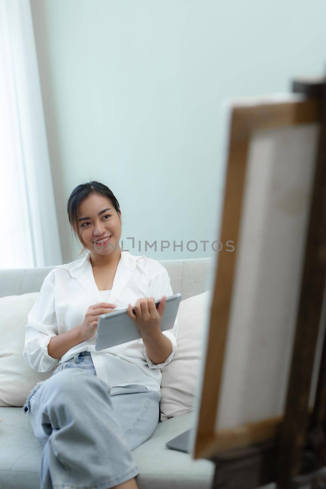 Portrait of an Asian woman using a tablet to paint artwork on a sofa. by Manastrong