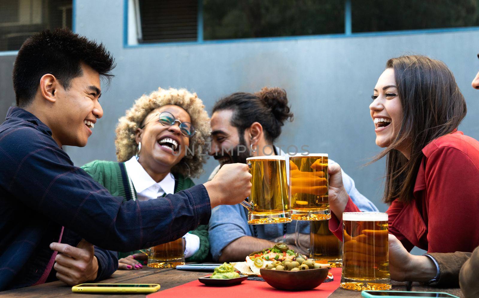 Cheers. Multiracial group of happy and smiling friends toast with beer and laugh together in a bar outdoors. by Hoverstock