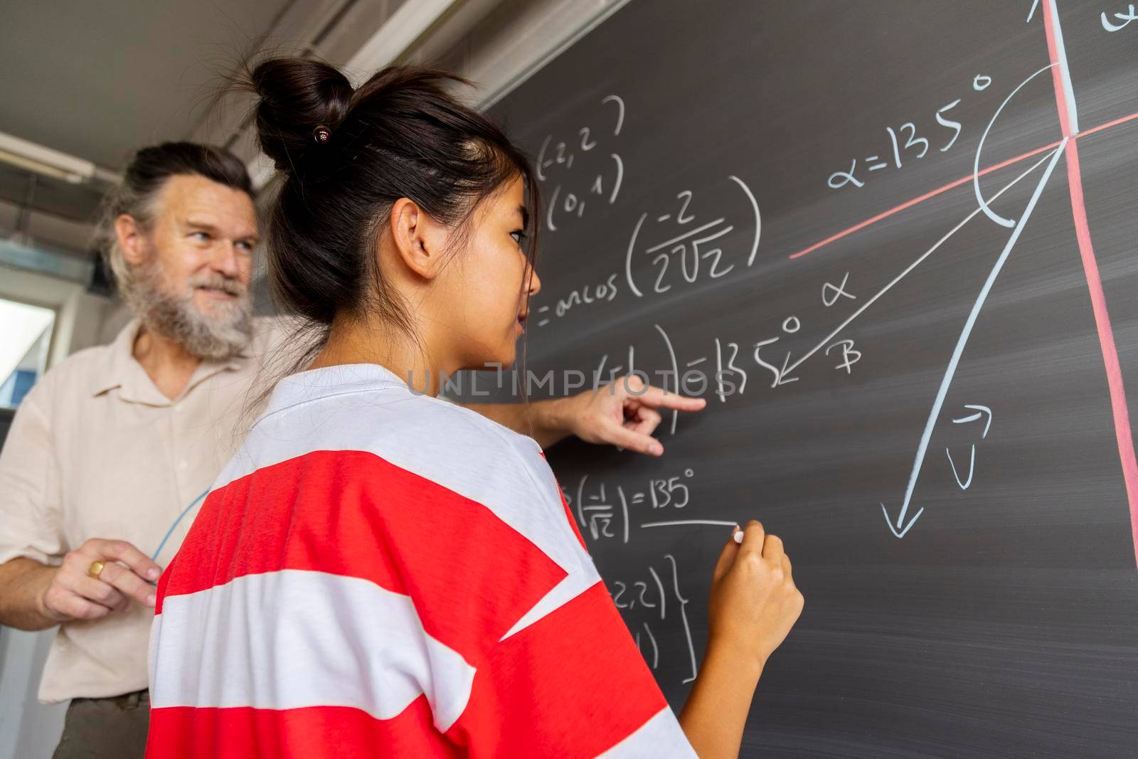 Mature caucasian male high school maths teacher explains blackboard exercise to asian girl teen student. by Hoverstock