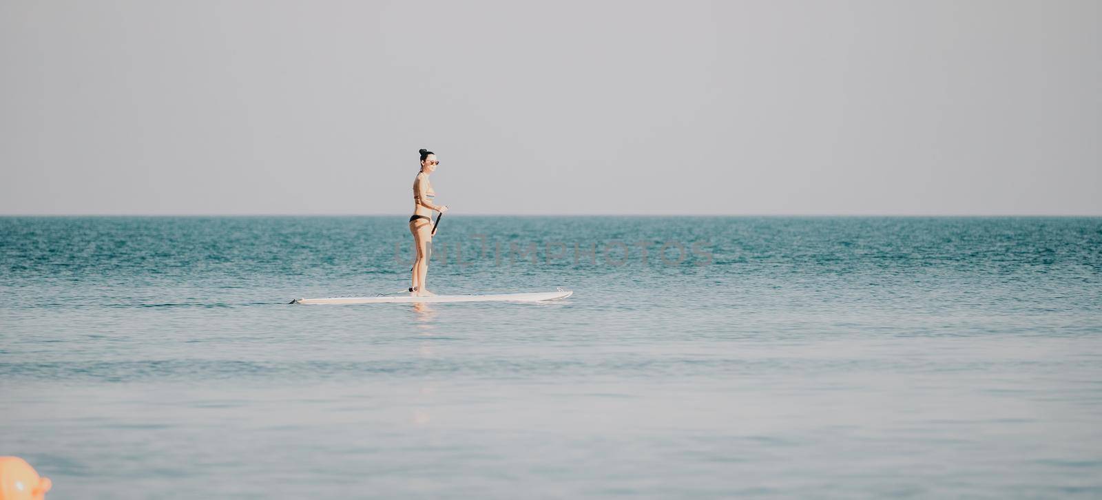 Silhouette of woman standing, surfing on SUP board, confident paddling through water surface. Idyllic sunset or sunrise. Sports active lifestyle at sea or river.