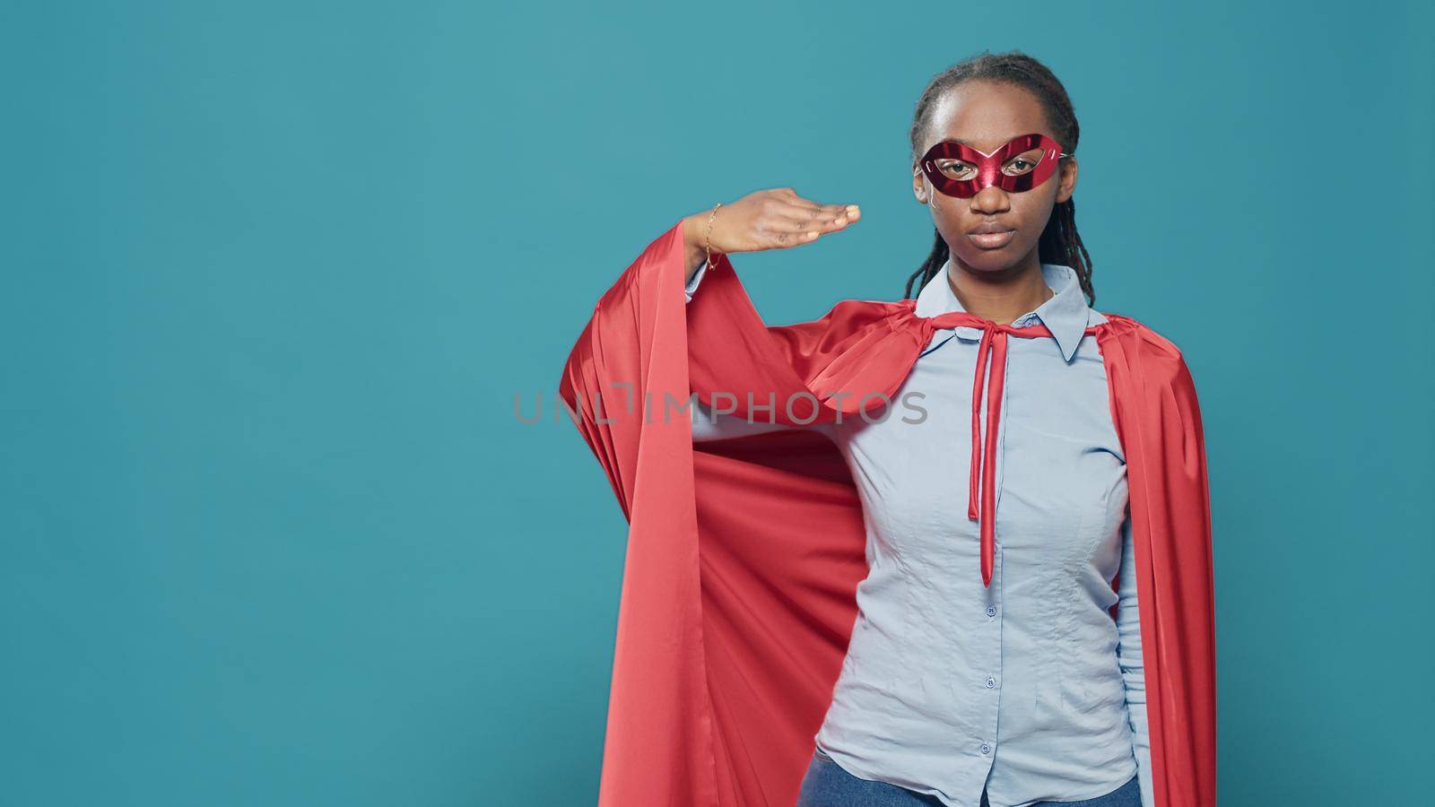 Portrait of woman superhero doing military salute to show motivation and respect. Wearing hero costume with red cape and mask, feeling confident and powerful over blue background in studio.
