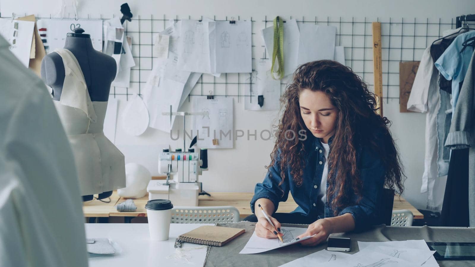 Young woman fashion designer is drawing ladies' garment sketch at desk in modern workshop. Mannequin, sewing items, to-go coffee are visible. Creativity in clothing manufacturing concept.