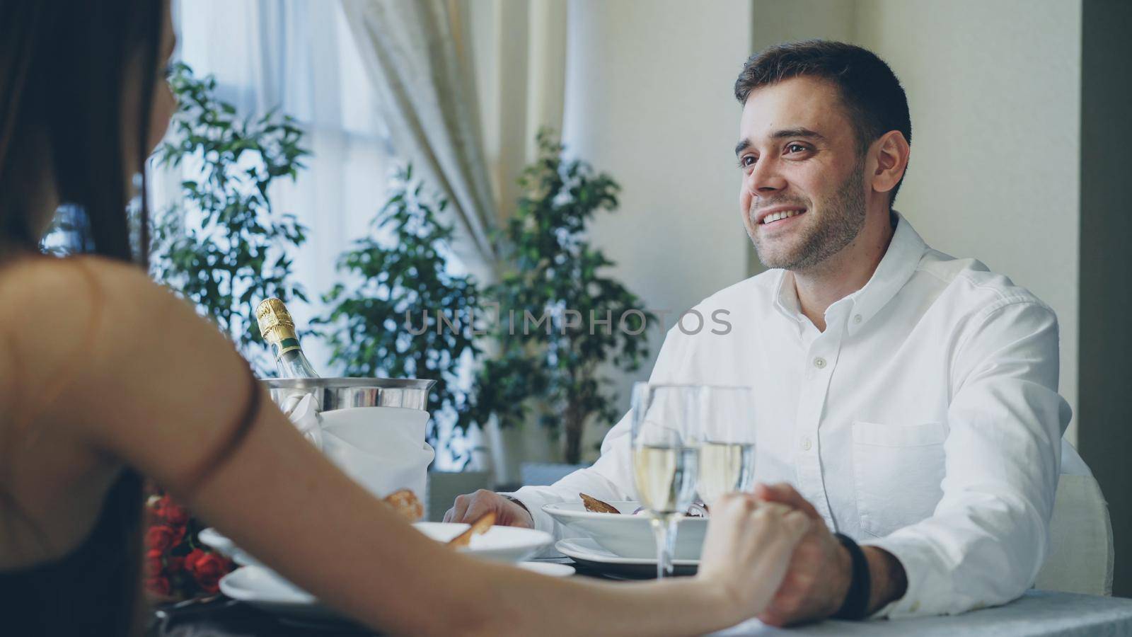 Young handsome man is talking to his girlfriend on date in fancy restaurant, drinking champagne and taking her hand with love and care. Romance and relationships concept.