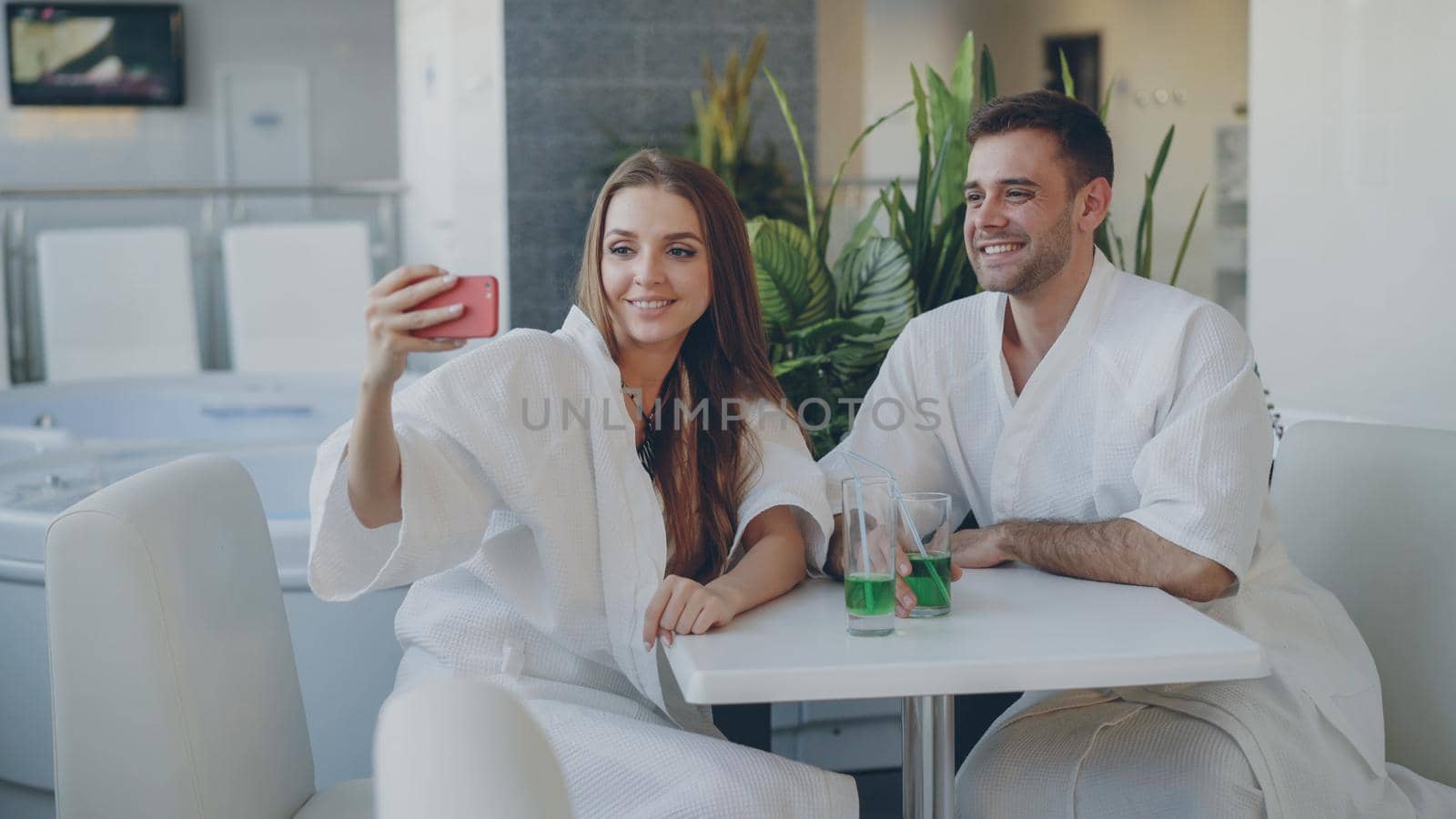 Pretty girl and her loving boyfriend are taking selfie with cocktail glasses using smartphone while relaxing in spa salon. They are smiling and posing looking at camera. by silverkblack