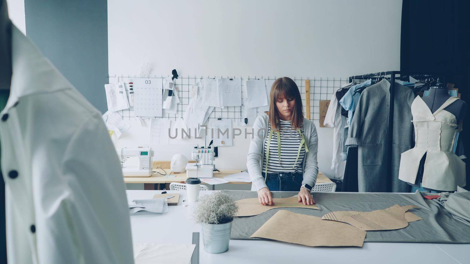 Attractive seamstress is measuring clothing paper templates lying on studio table with measure-tape. Woman is calm and concentrated on work in light tailor's shop.