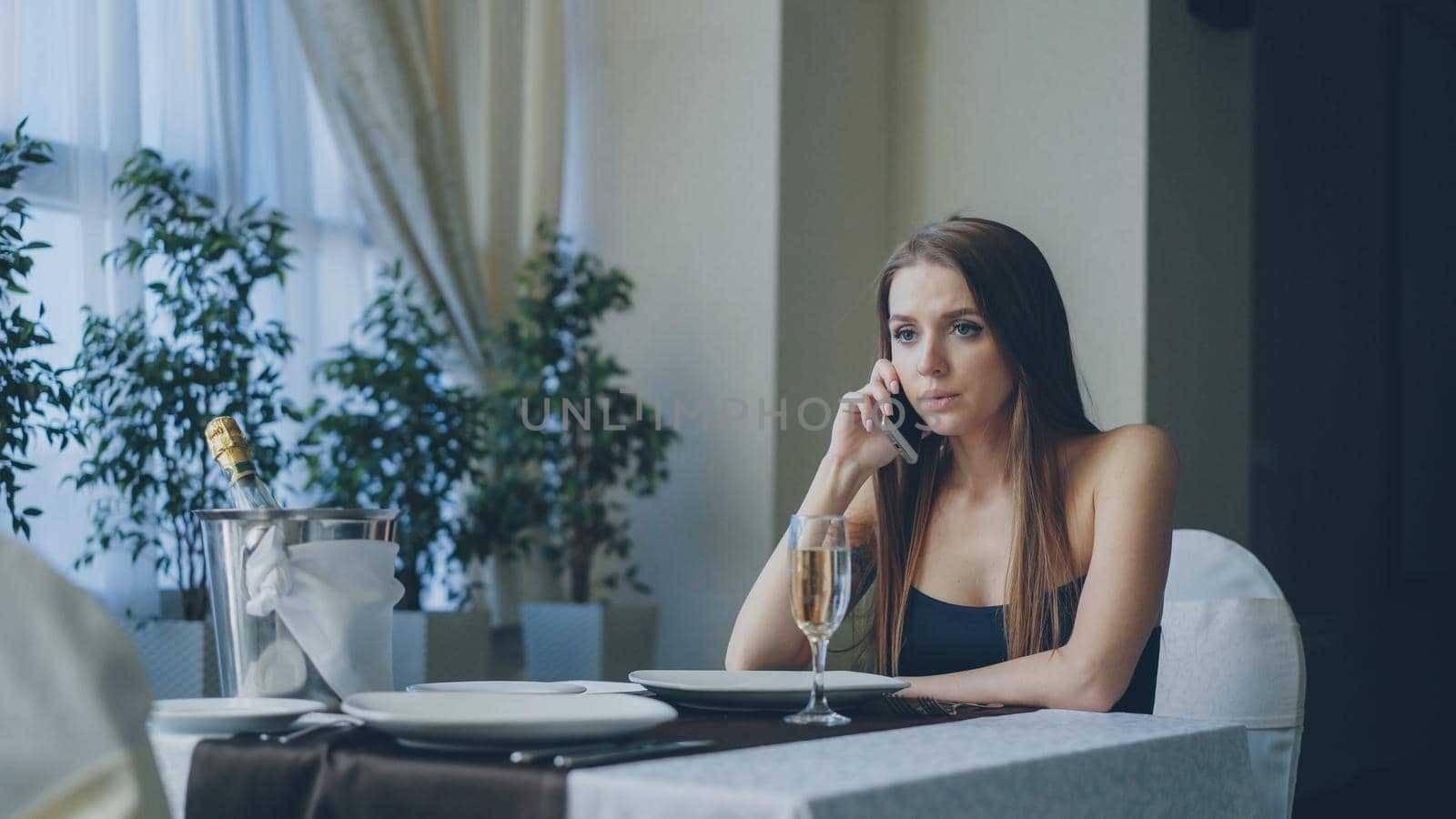 Angry beautiful woman is calling her boyfriend on mobile phone while waiting for him alone in restaurant. Girl is impatient, nervous and offended. by silverkblack