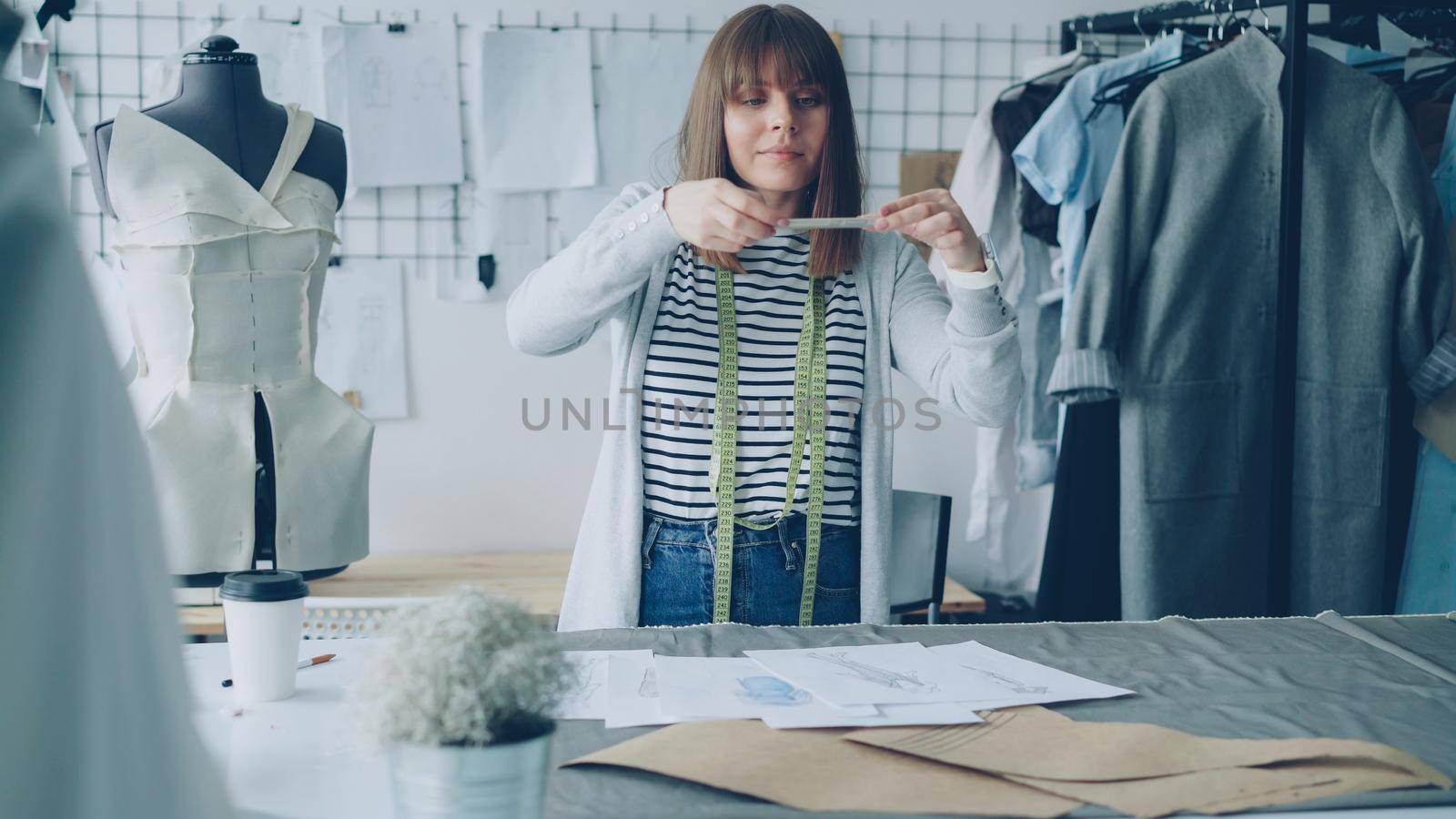 Beautiful young woman fashion designer is taking photos of sketches lying on table. She is making creative flat lays. Her studio is light and full of sewing items, tools and equipment.