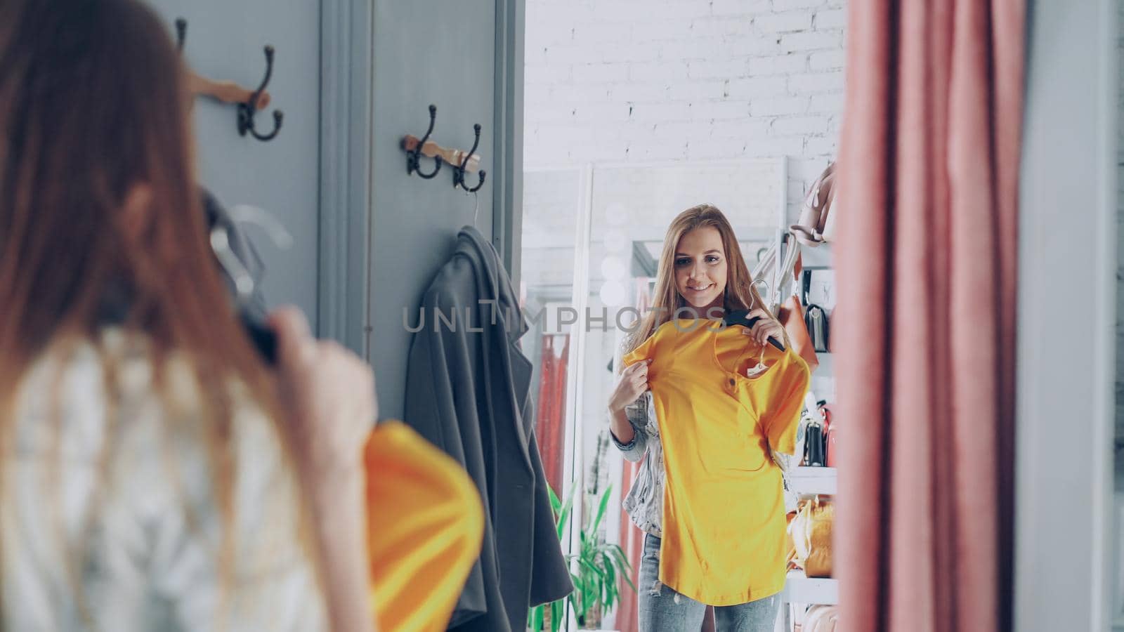 Mirror shot of young lady choosing clothes in fitting room. Girl is trying top checking size and length while standing opposite large mirror. Nice clothing store in background.