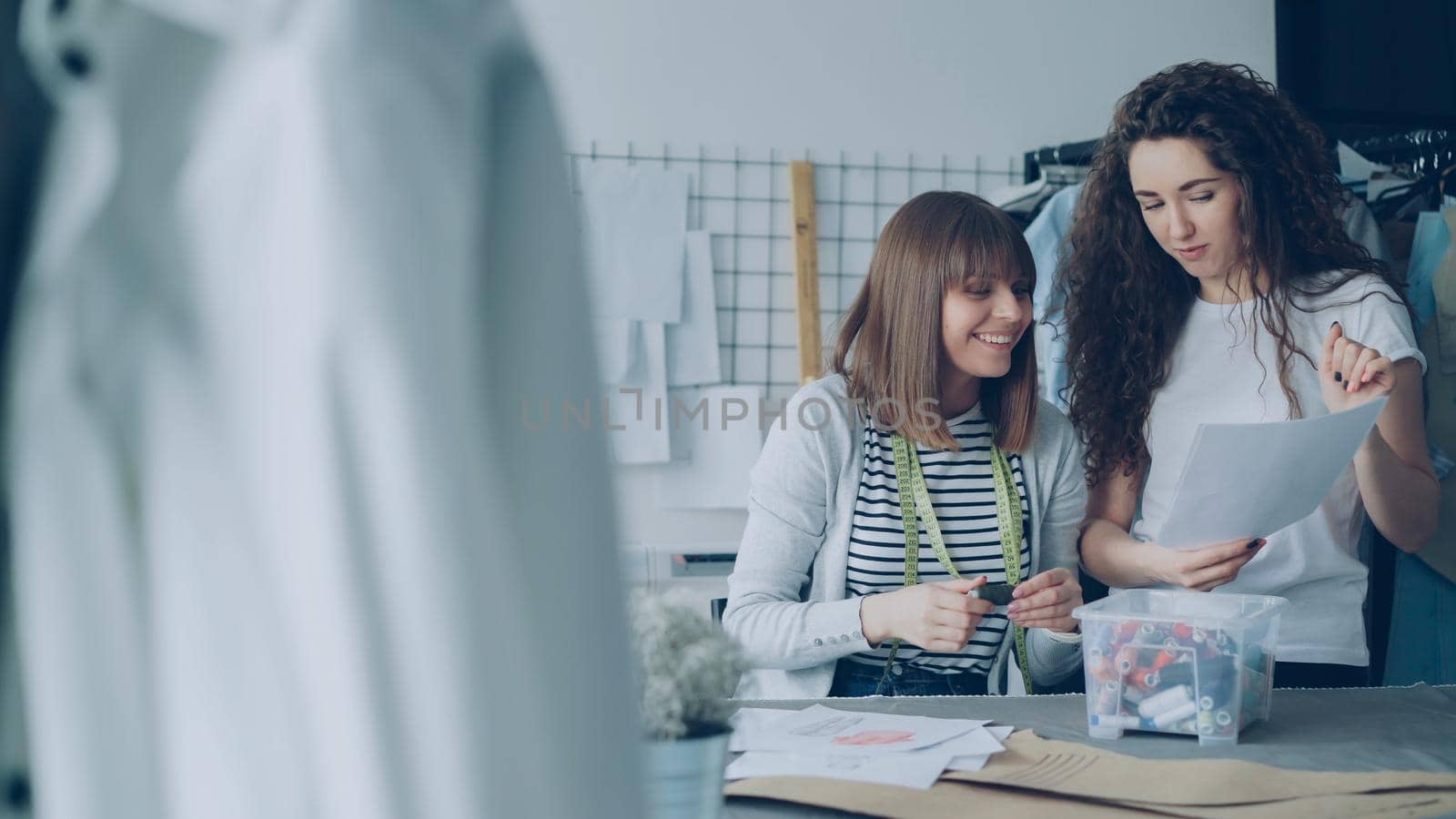 Female sewer is reeling in and out colofrul sewing threads while her coworker is bringing garment sketches and showing them to her. Women are choosing colors and discussing future clothes.