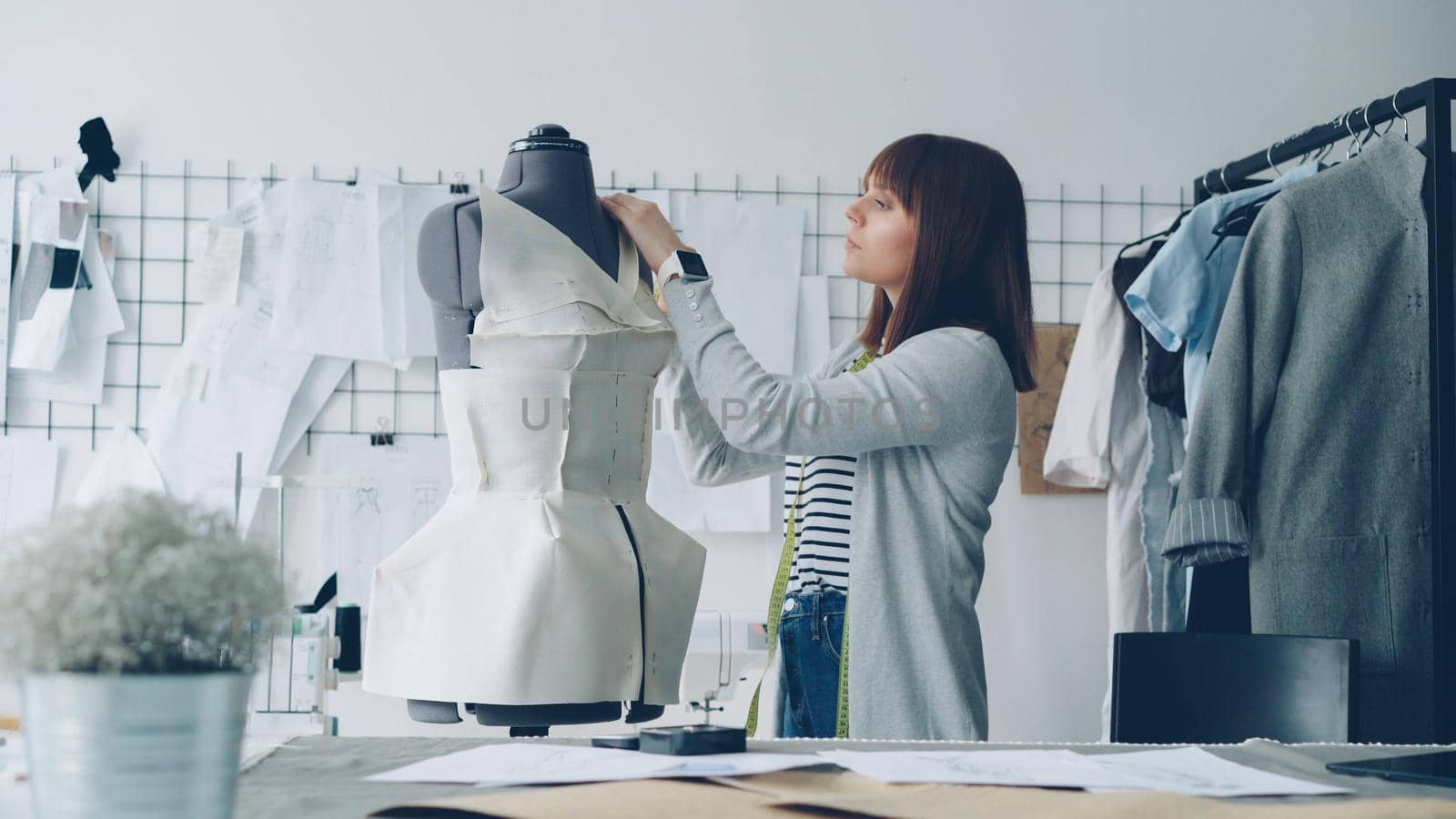 Young attractive seamstress is measuring tailor's dummy with tape to make new garment with these measurements. Busy day in clothing workshop concept. by silverkblack