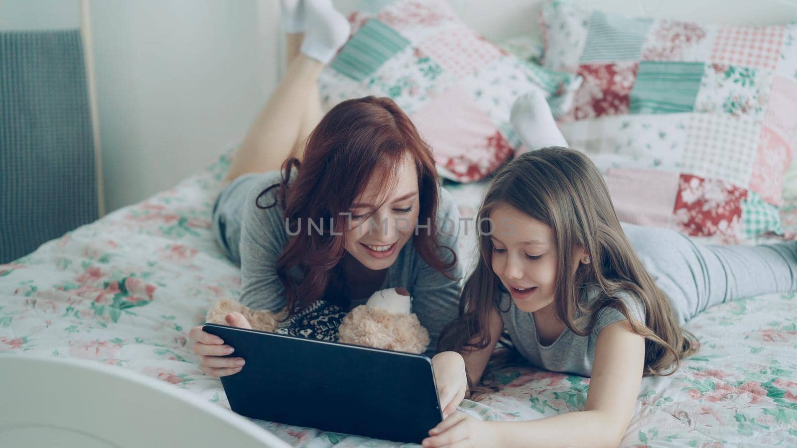 Beautiful young mother and her cute daughter in pajamas laughing and looking in digital tablet while lying on bed at home in the morning by silverkblack