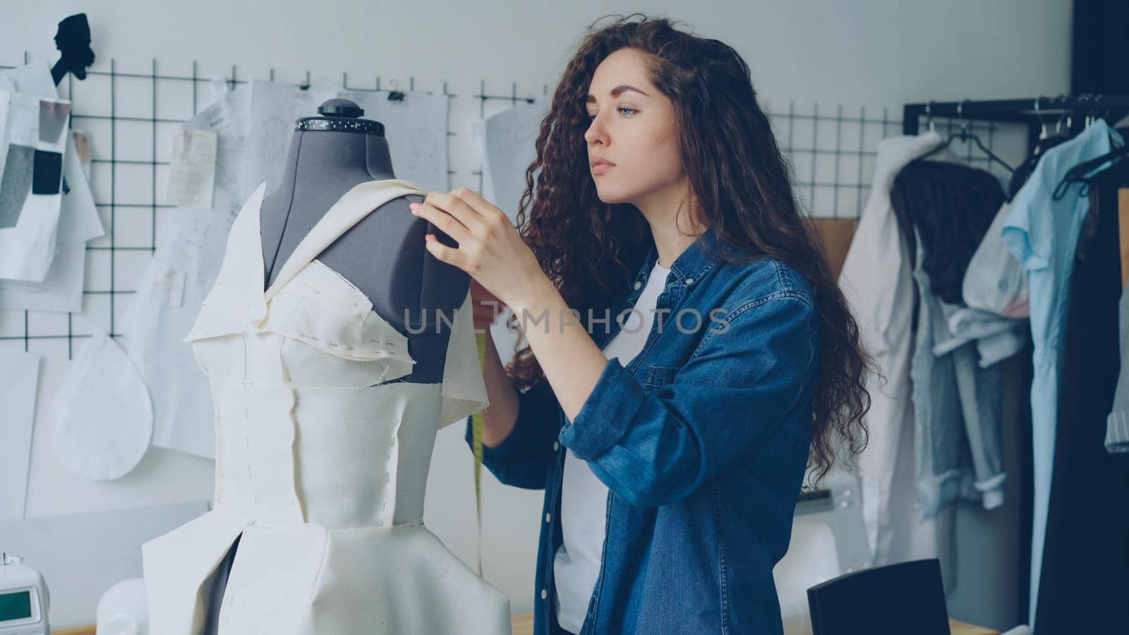 Young female tailor is adjusting clothes on tailoring dummy with sewing pins and measuring with measure-tape. Women's garments, sketches on wall, tailoring items and tools are visible.