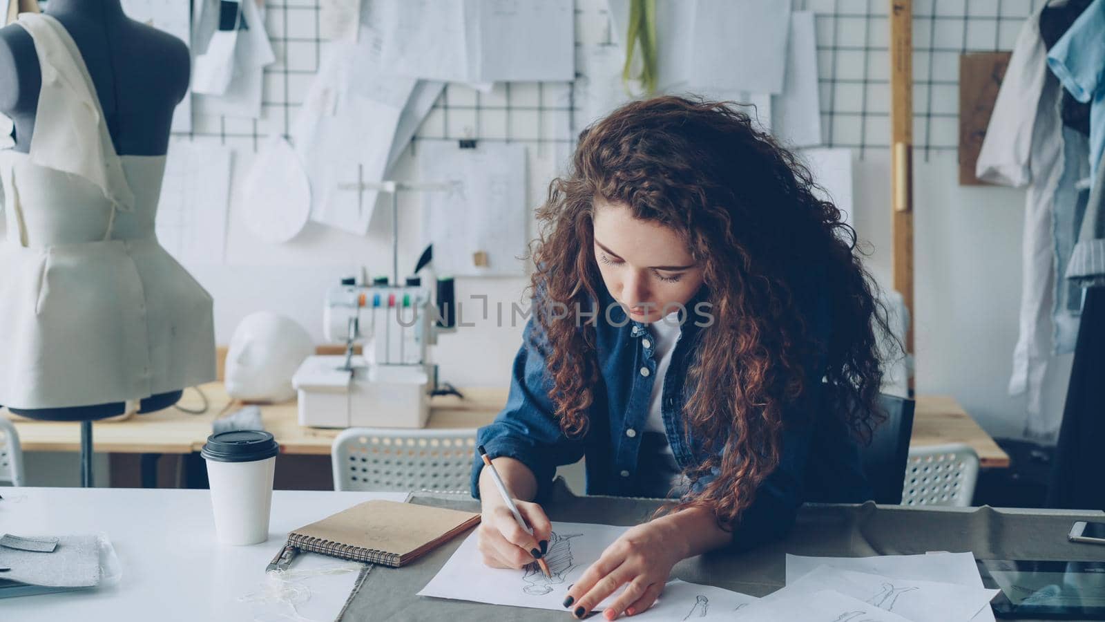 Young female artist fashion designer is drawing women's garment sketch at table in modern studio. Mannequin, sewing items, take-away coffee are visible. by silverkblack