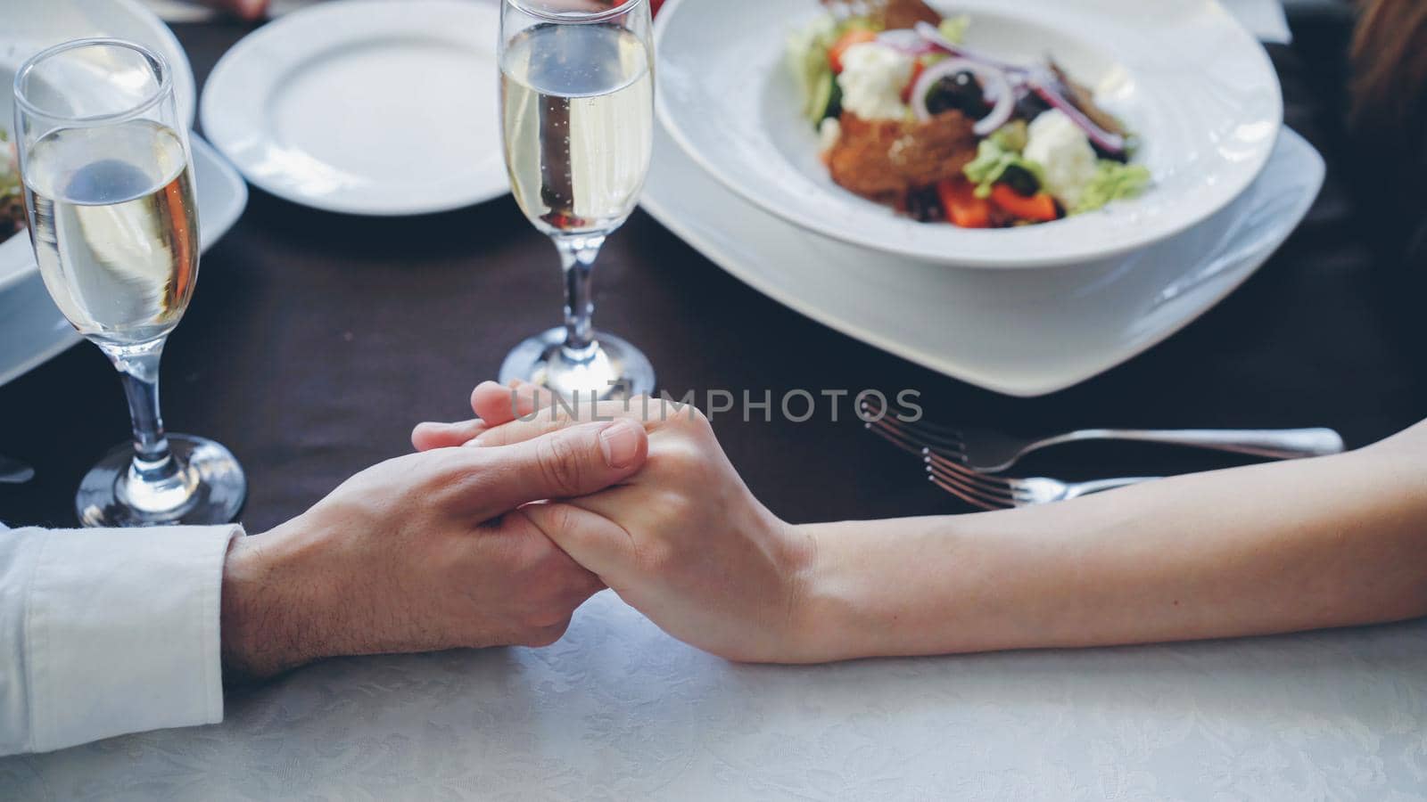 Close-up shot of young lovers touching and holding hands at romantic dinner in classy restaurant. Table with sparking champagne glasses, flatware and food in background.