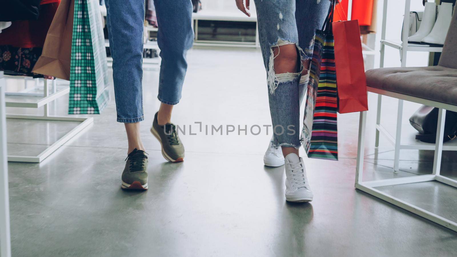 women's legs walking slowly through luxurious shop. Women are wearing jeans and trainers and carrying bright paper bags. by silverkblack