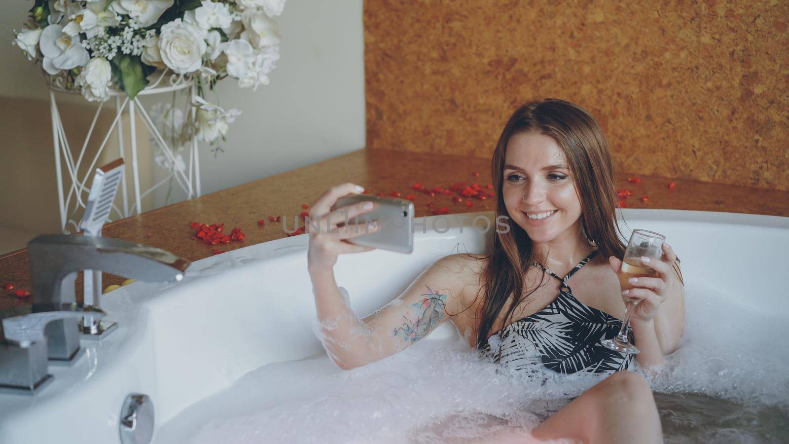 Pretty young woman is taking selfie with champagne glass using smartphone in hot tub in modern spa salon. She is smiling and posing looking at camera. by silverkblack