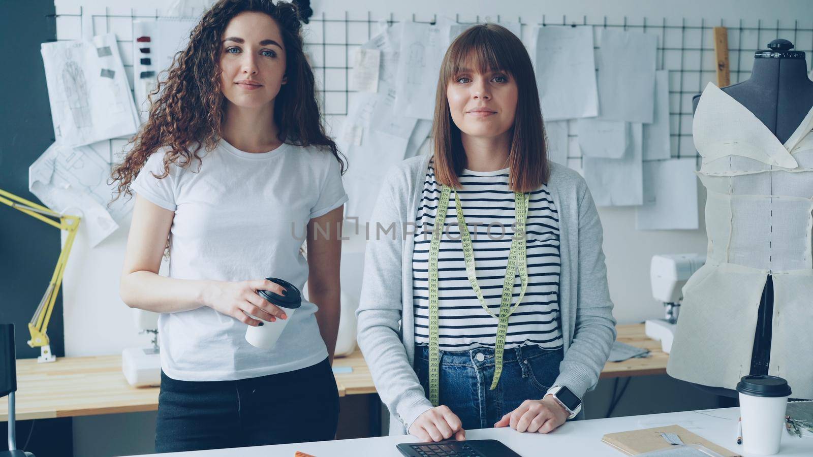 Portrait of two female clothing designers entrepreneurs standing in workshop together and looking at camera. One woman is holding take-out coffee, other is touching studio desk. by silverkblack