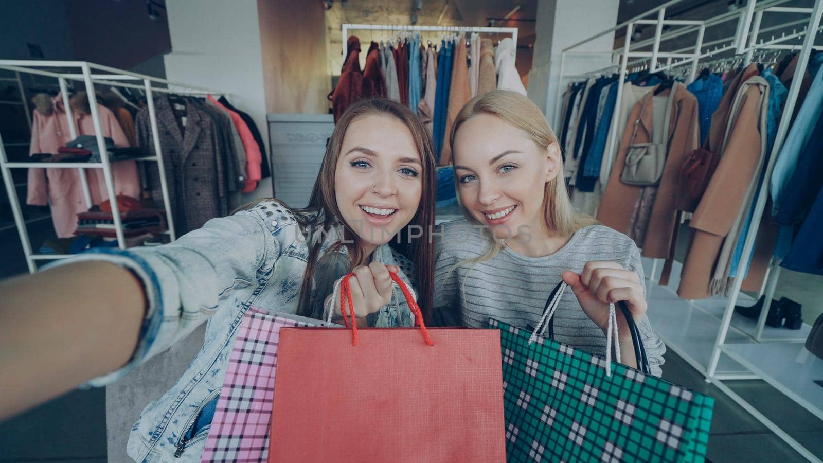 Point of view shot of two attractive careless girls making selfie with paper bags in women's clothes shop. Friends are posing, laughing and chatting happily by silverkblack