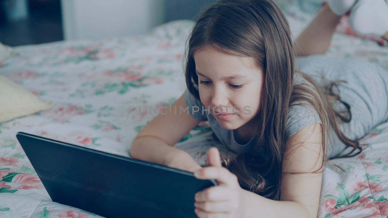 Closeup of Cute little girl using digital tablet and smiling while lying in bed. Child wathcing cartoon movie on portable device and laughing at home by silverkblack