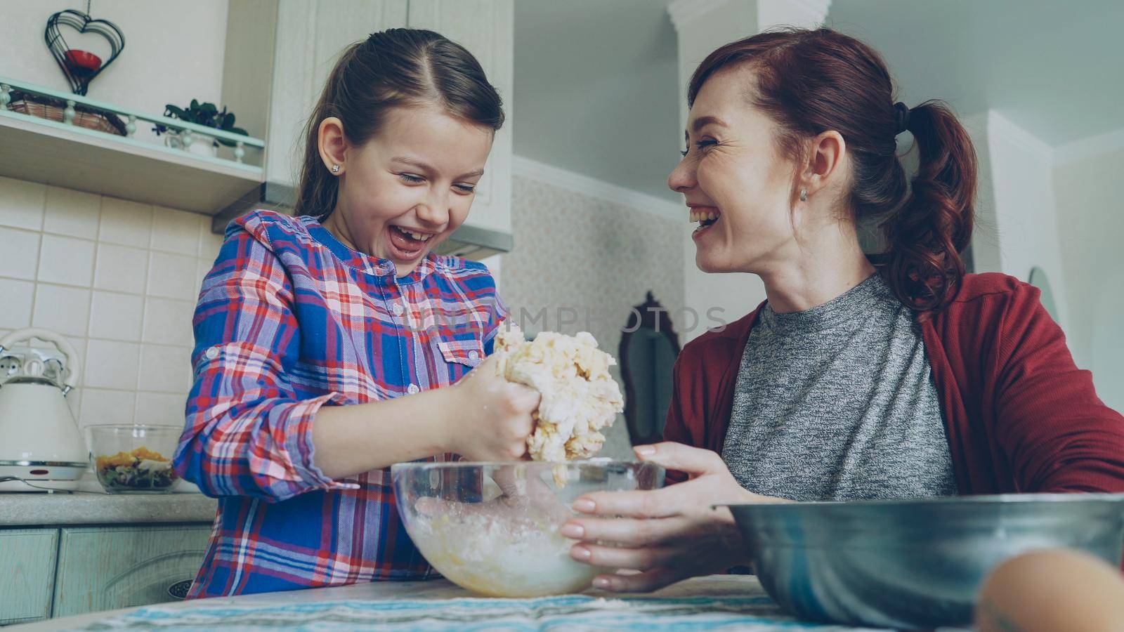 Little pretty girl helping her mother in the kitchen stirring dough for cookies into bowl. Mom and daughter have fun talking and laughing. Family, food, home and people concept