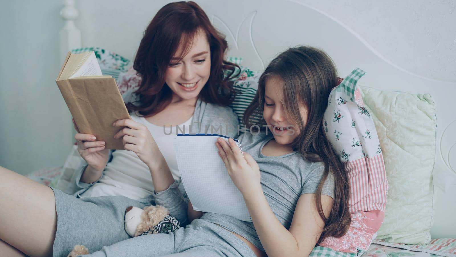 Careful mother helping her little cute daughter with homework for elementary school. Loving mom reading a book and girl writing notes in copybook while sitting together on bed at home by silverkblack