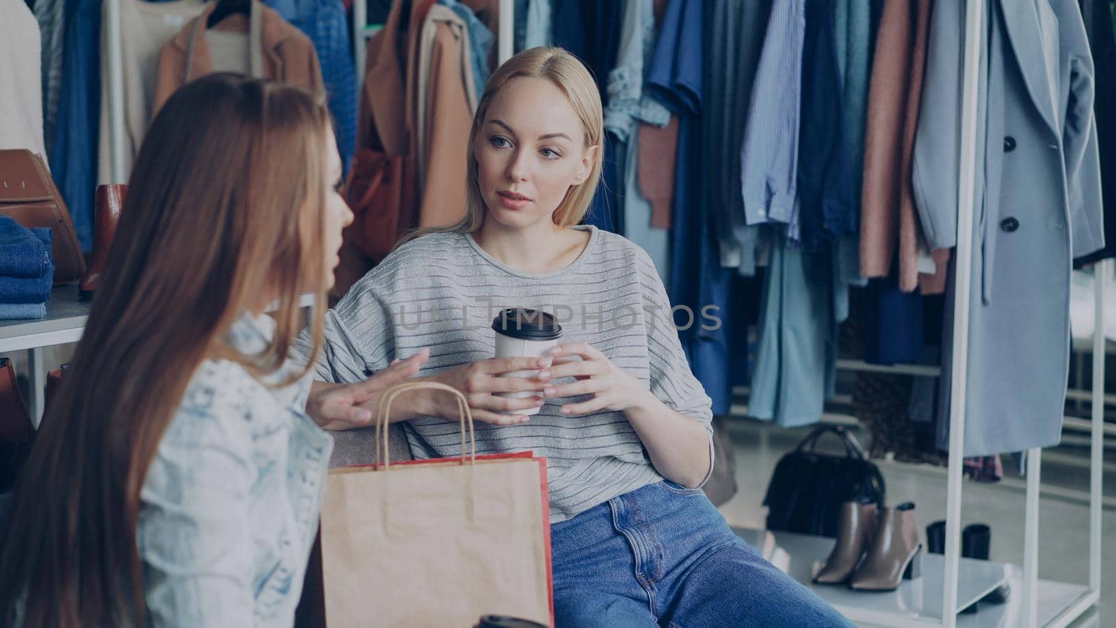 Young attractive woman sitting in modern clothes shop, holding coffee and talking to her female friend. Ladies having rest after going shopping.