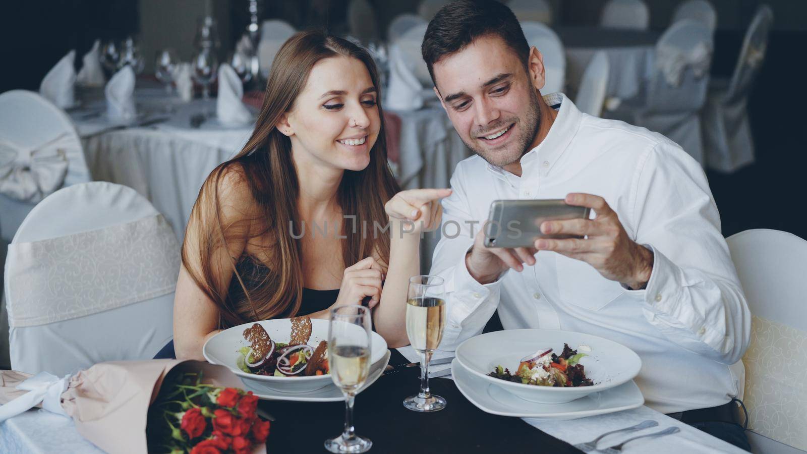 Happy attractive young people are watching smartphone together, smiling and talking while having romantic dinner in restaurant. Modern technologies and romance concept.