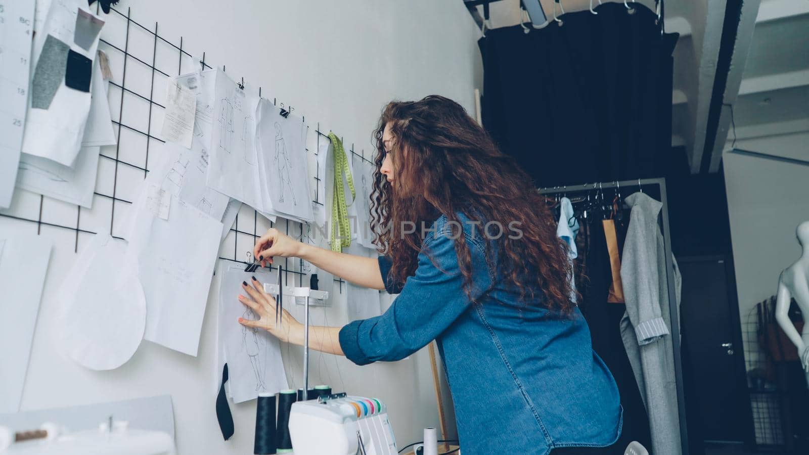 Low angle view of young attractive fashion designer looking at sketches and hanging drawings on wall in modern loft style studio. Large collection of illustration above tailoring desk.