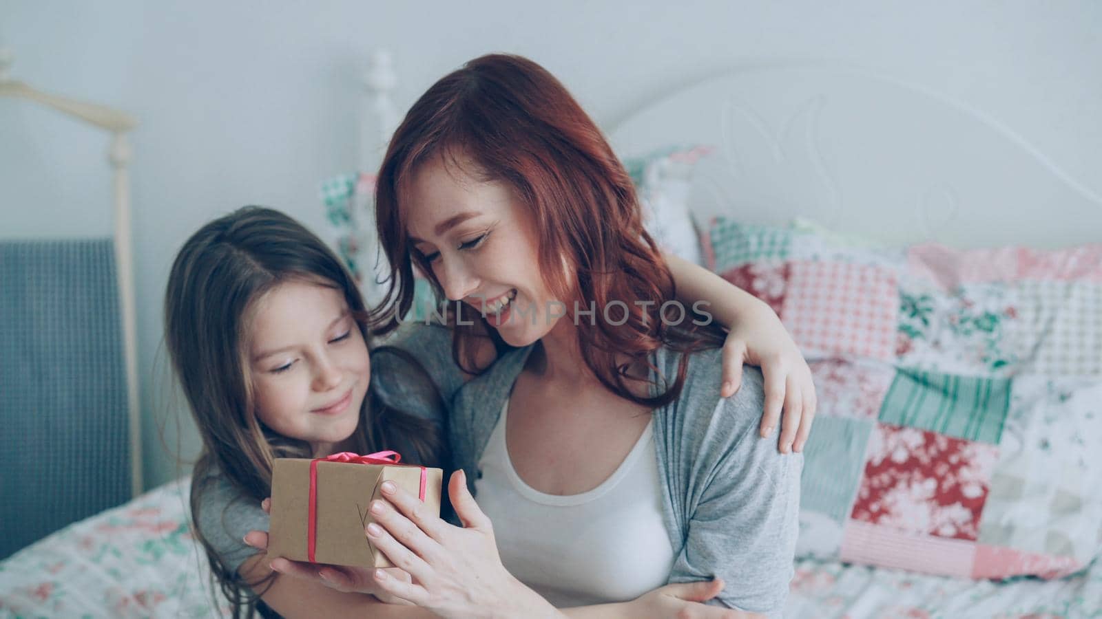 Little cute girl giving gift box to her young happy mom celebrating mothers day sitting on bed in bright bedroom at home