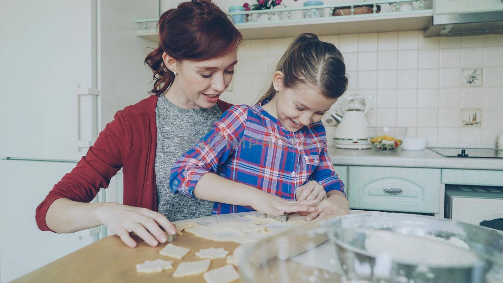Cheerful mother and little cute daughter talking and making cookies together using bakery forms cropping dough while sitting in modern kitchen at home. Family, food and people concept by silverkblack