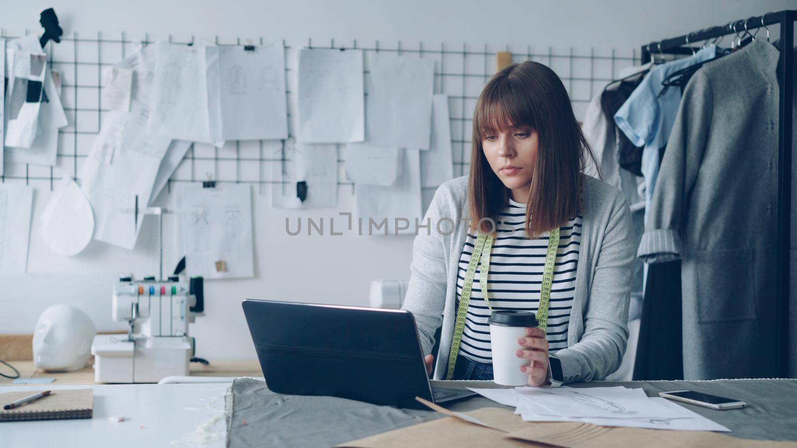 Young attractive seamstress is working with laptop and drinking take-out coffee in her tailoring studio. She is having rest after work. Relaxation in workplace concept.