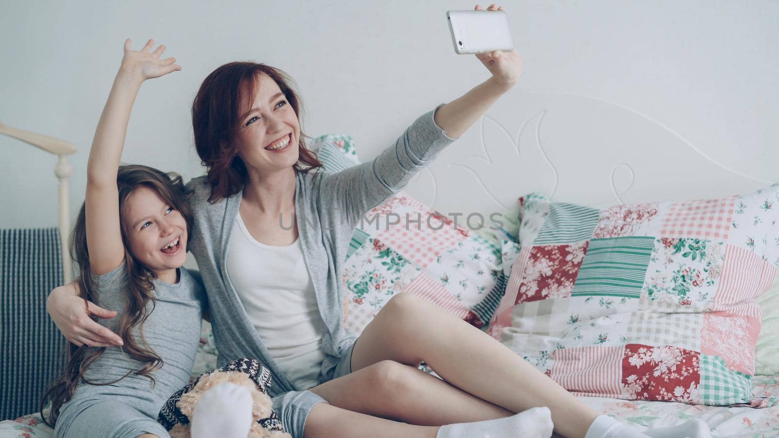 Happy mother and little girl taking selfie photo with smartphone camera and have fun grimacing while sitting in cozy bedroom at home