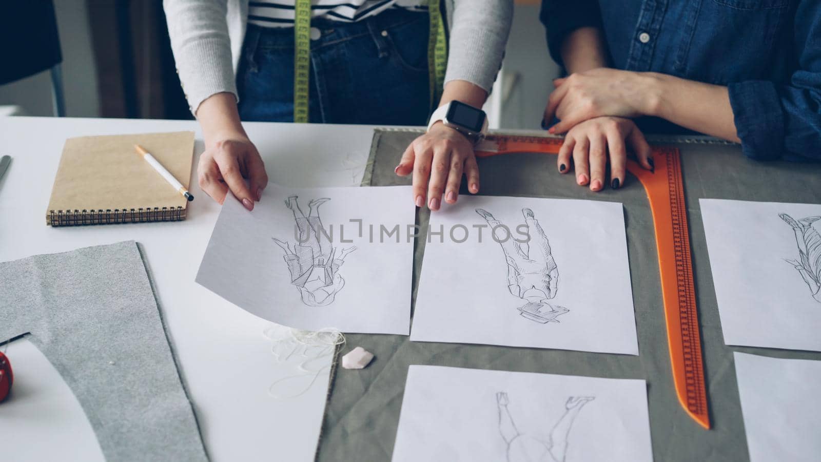 Close-up shot of tailors' manicured hands going through black-and-white sketches on sewing desk. Young women are discussing clothing designs. Creative dressmaker's work concept.