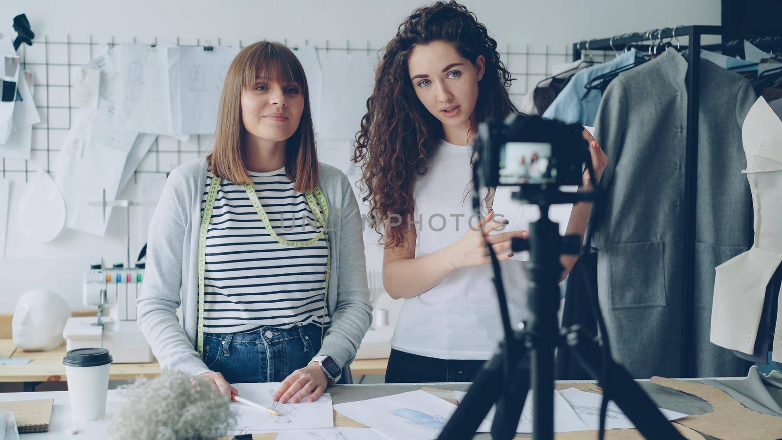 Young pretty women fashion designers bloggers are recording video with camera about sewing new garment. Girls are showing sketches and templates and talking together. by silverkblack