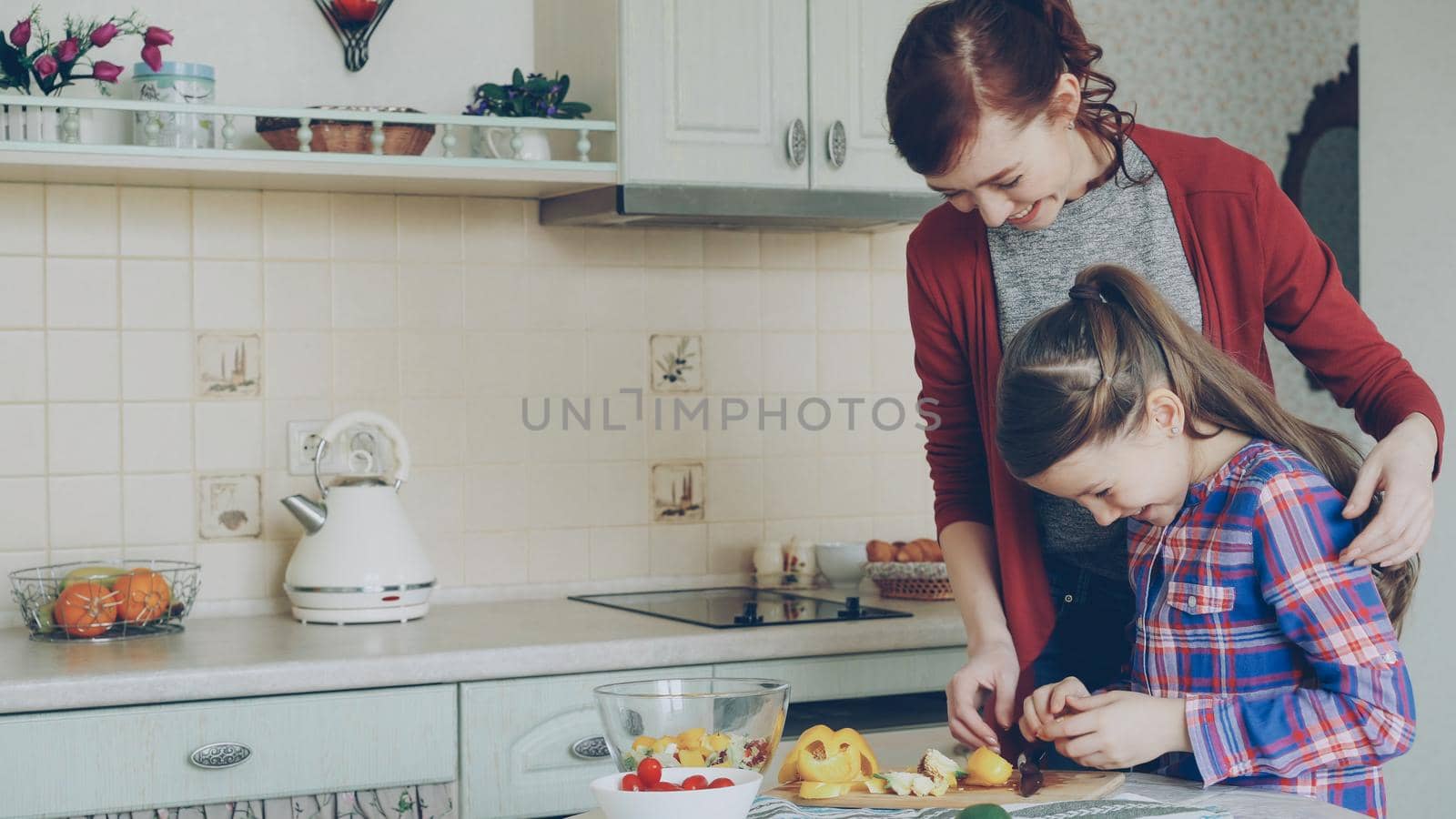 Young pretty mother teaching her cute daughter to cut vegetables properly. Little girl cooking together with loving mom at home in modern kithcen by silverkblack