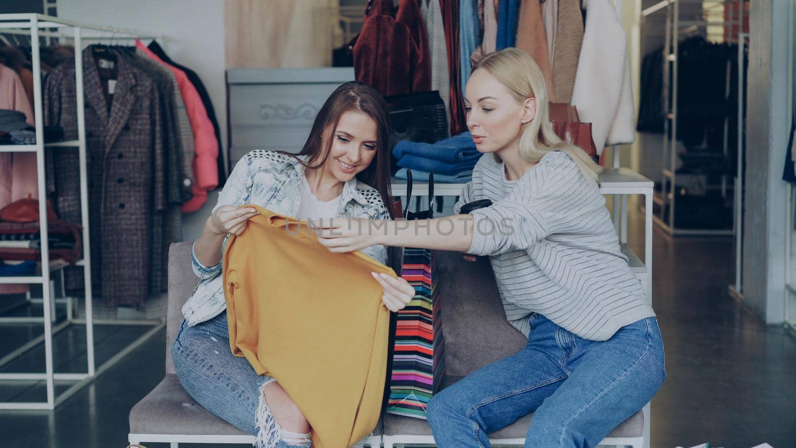 Happy young woman telling her friend about newly purchased knitted jumper, then showing it to her while sitting in shop. Female friend touching it, checking quality and approving her choice.