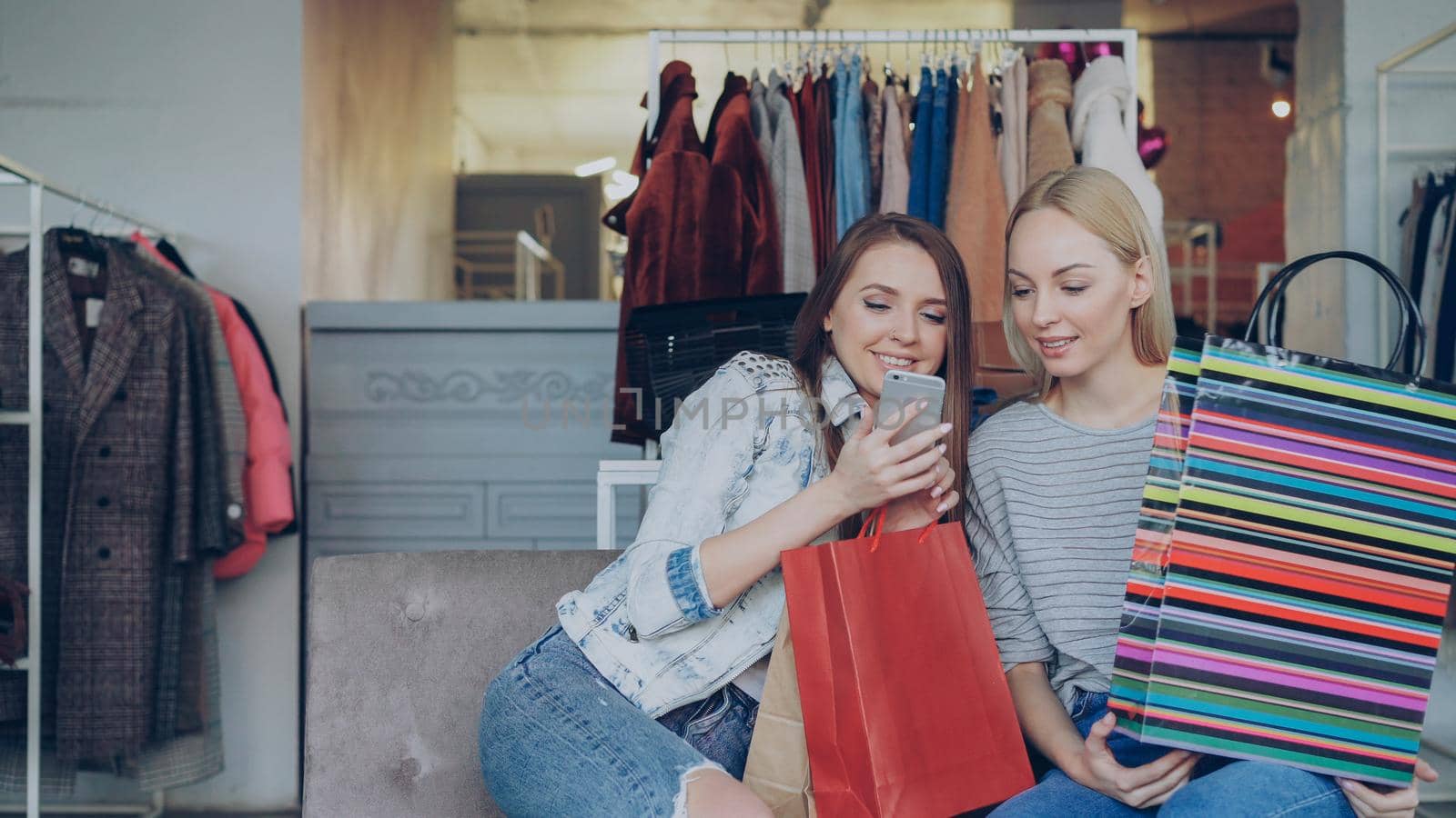 Cheerful young women chatting and using modern smartphone while sitting in nice clothing boutique. They are smiling and gesturing enthusiastically. by silverkblack