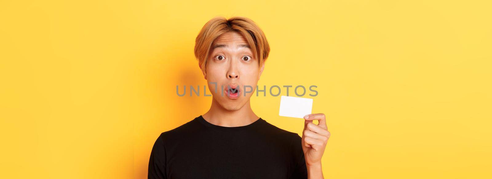 Close-up of surprised gasping asian guy with fair hair, looking amazed and startled as showing credit card, standing yellow background.