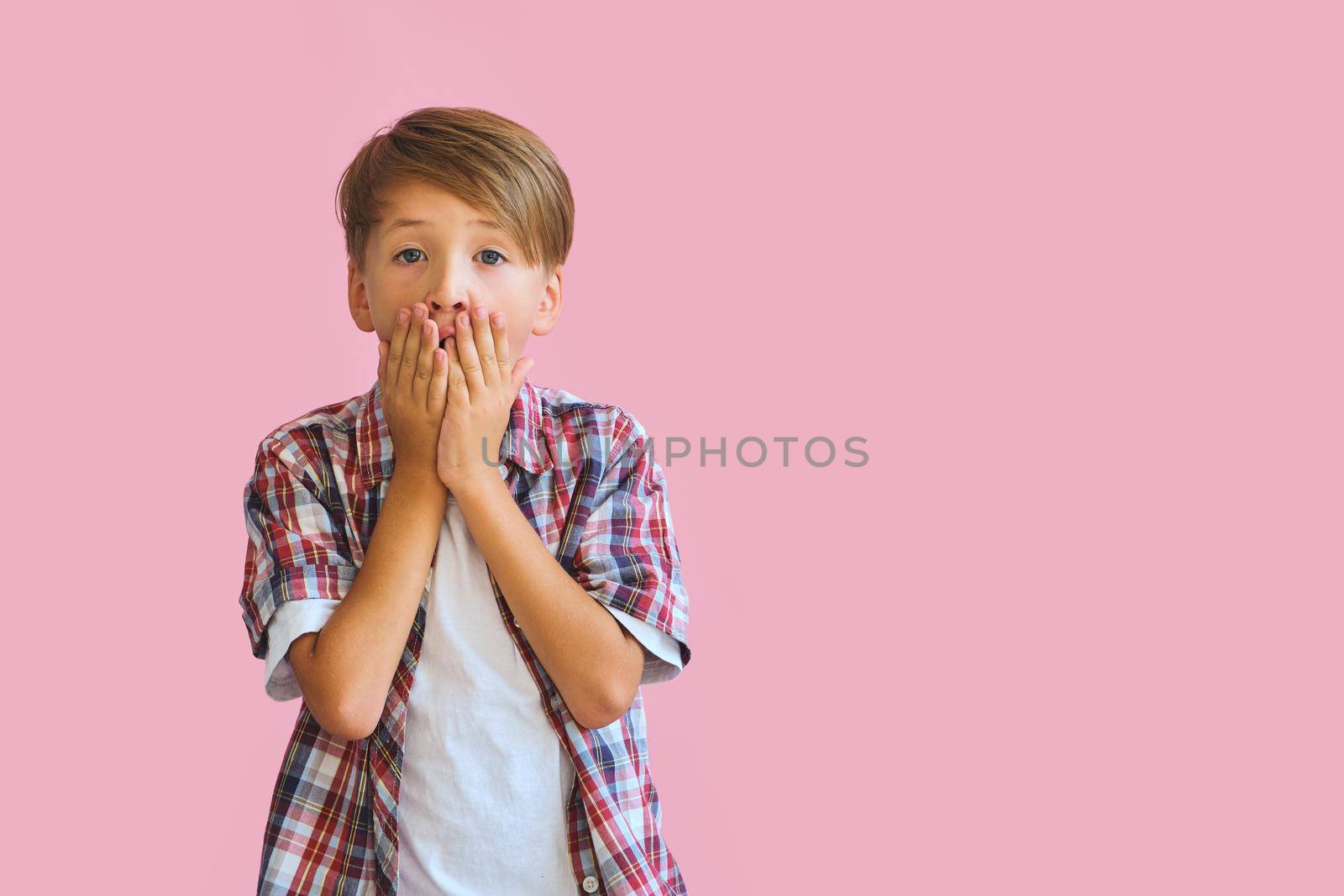 Young surprised boy dressed in jeans, a white T-shirt and a plaid shirt covers mouth with palms, isolated on pink background with copy space